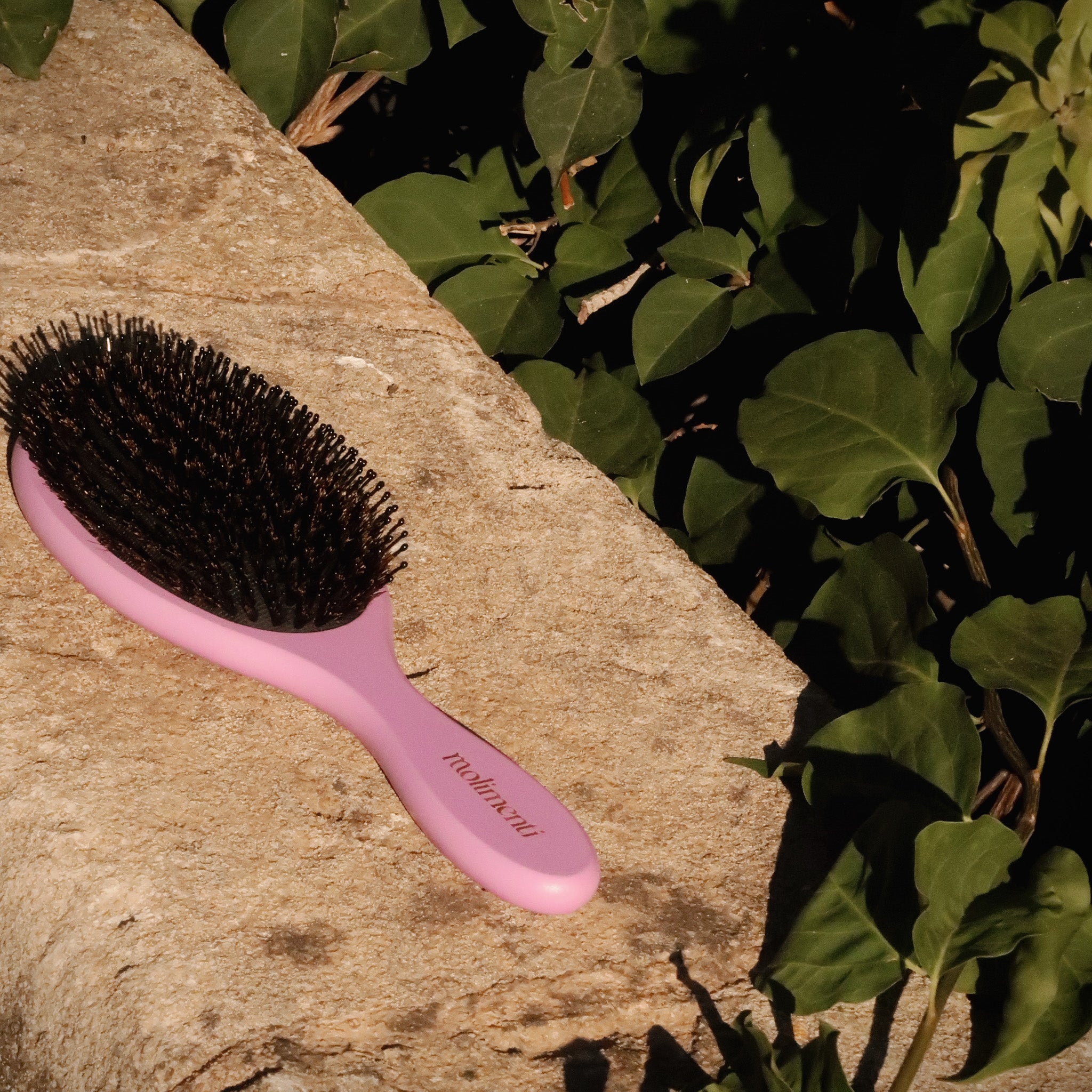 Your Hair Brush – Lavender Large från Molimenti, liggande på en solvarm stenmur med grönska i bakgrunden. Den lila hårborsten med svart borsthuvud fångas i varmt dagsljus för en naturlig och somrig känsla.