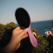 Your Hair Brush – Lavender Large från Molimenti, hålls upp framför en havsutsikt med båtar och grönska i bakgrunden. Lila borste med svart borsthuvud i naturligt ljus, fotograferad i en somrig medelhavsmiljö.