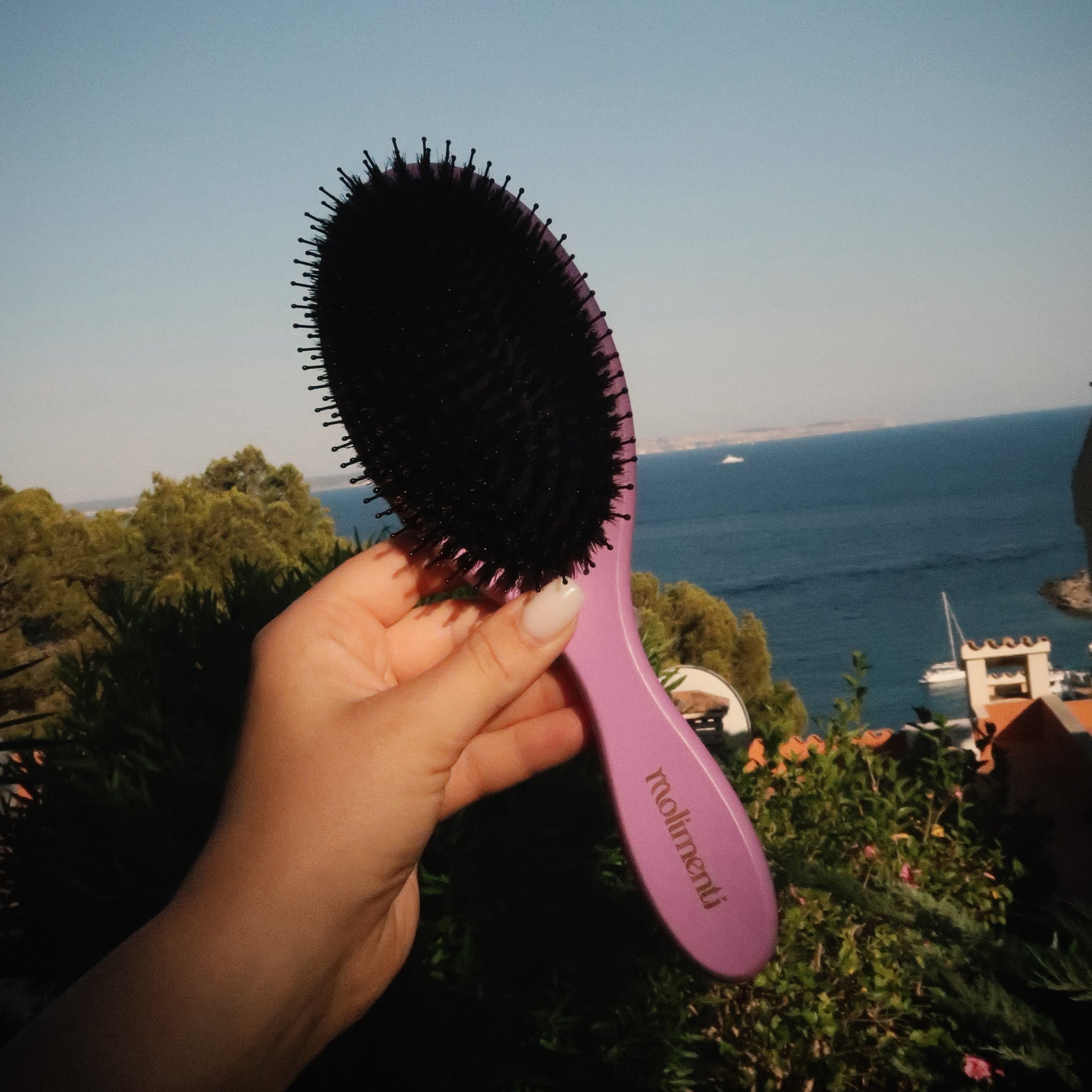 Your Hair Brush – Lavender Large från Molimenti, hålls upp framför en havsutsikt med båtar och grönska i bakgrunden. Lila borste med svart borsthuvud i naturligt ljus, fotograferad i en somrig medelhavsmiljö.