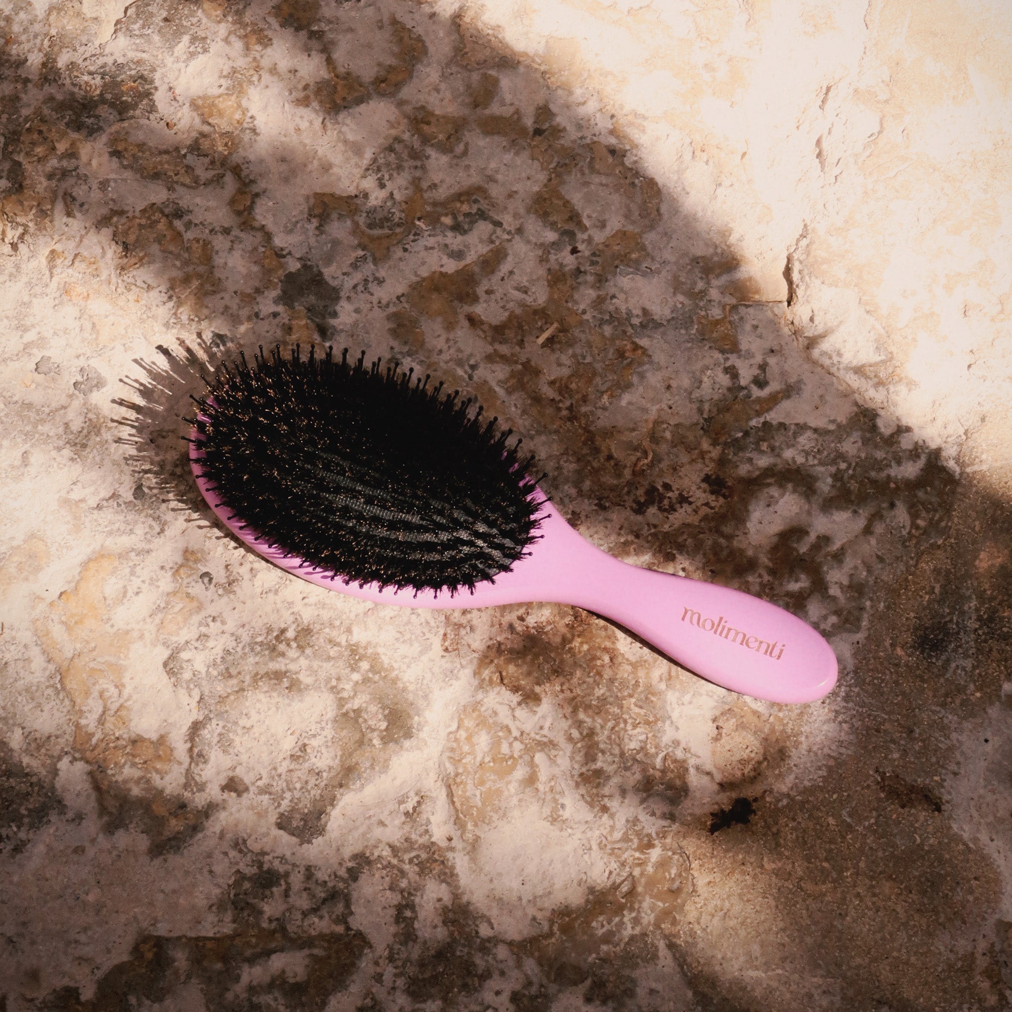 Your Hair Brush – Lavender Large från Molimenti, liggande på solbelyst natursten med varma skuggor. Den lila hårborsten med svart borsthuvud fångas i ett naturligt ljus som framhäver textur och färg.